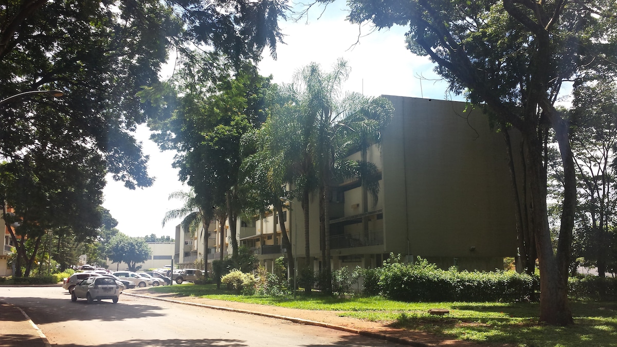 The building is set amidst lush greenery, with tall trees providing shade. Vehicles are parked along the side of the street, which leads to a well-maintained area indicating a tranquil residential environment. Sunlight filters through the branches, contributing to a calm atmosphere.