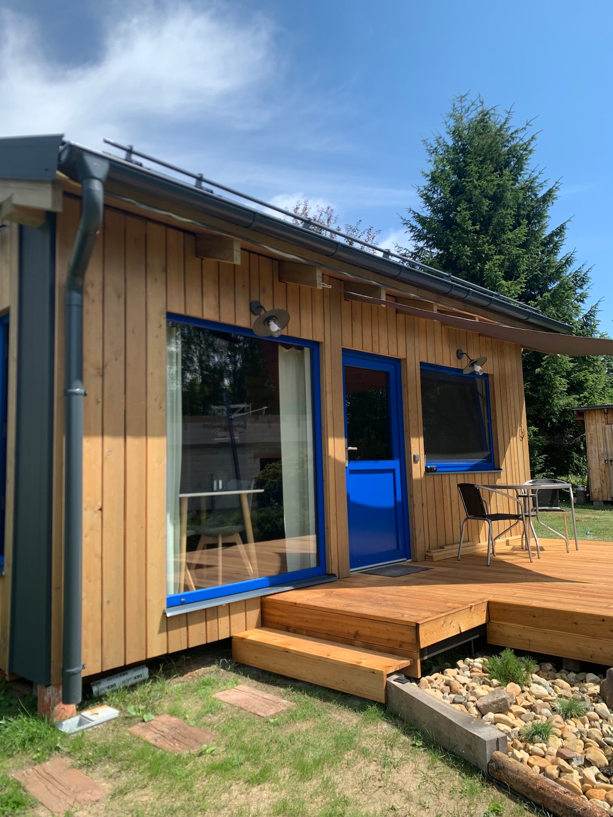 The exterior of the house is clad in wooden panels with blue trim, featuring large windows that allow natural light to fill the space. A wooden deck leads to a small table and chairs set, inviting guests to enjoy the outdoor area.