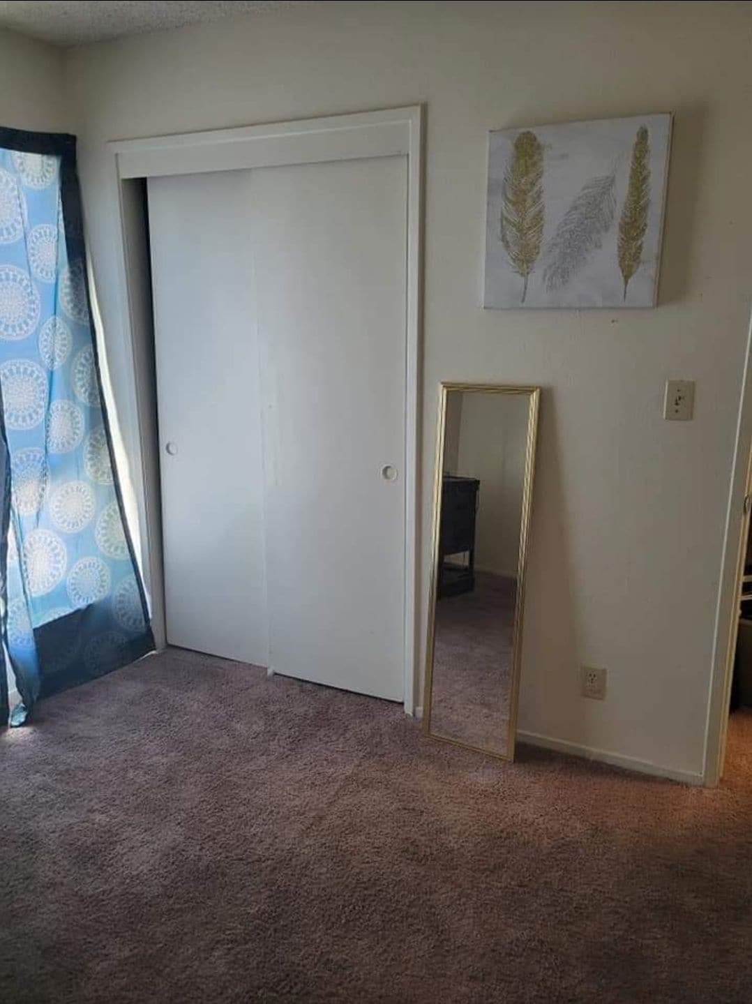 The room features a light-colored carpet and neutral walls. A full-length, gold-framed mirror stands against one wall. A closet with sliding doors is visible, and a window covered by a blue patterned curtain allows natural light to enter the space.