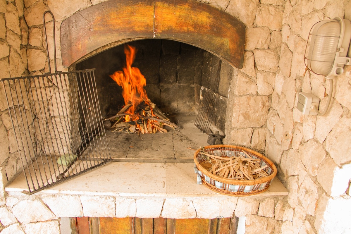 A rustic stone fireplace is featured, with a fire crackling within and flames visible. Adjacent, a woven basket filled with kindling sits on the stone hearth. The warm tones of the stones create a cozy atmosphere in the outdoor grilling area.