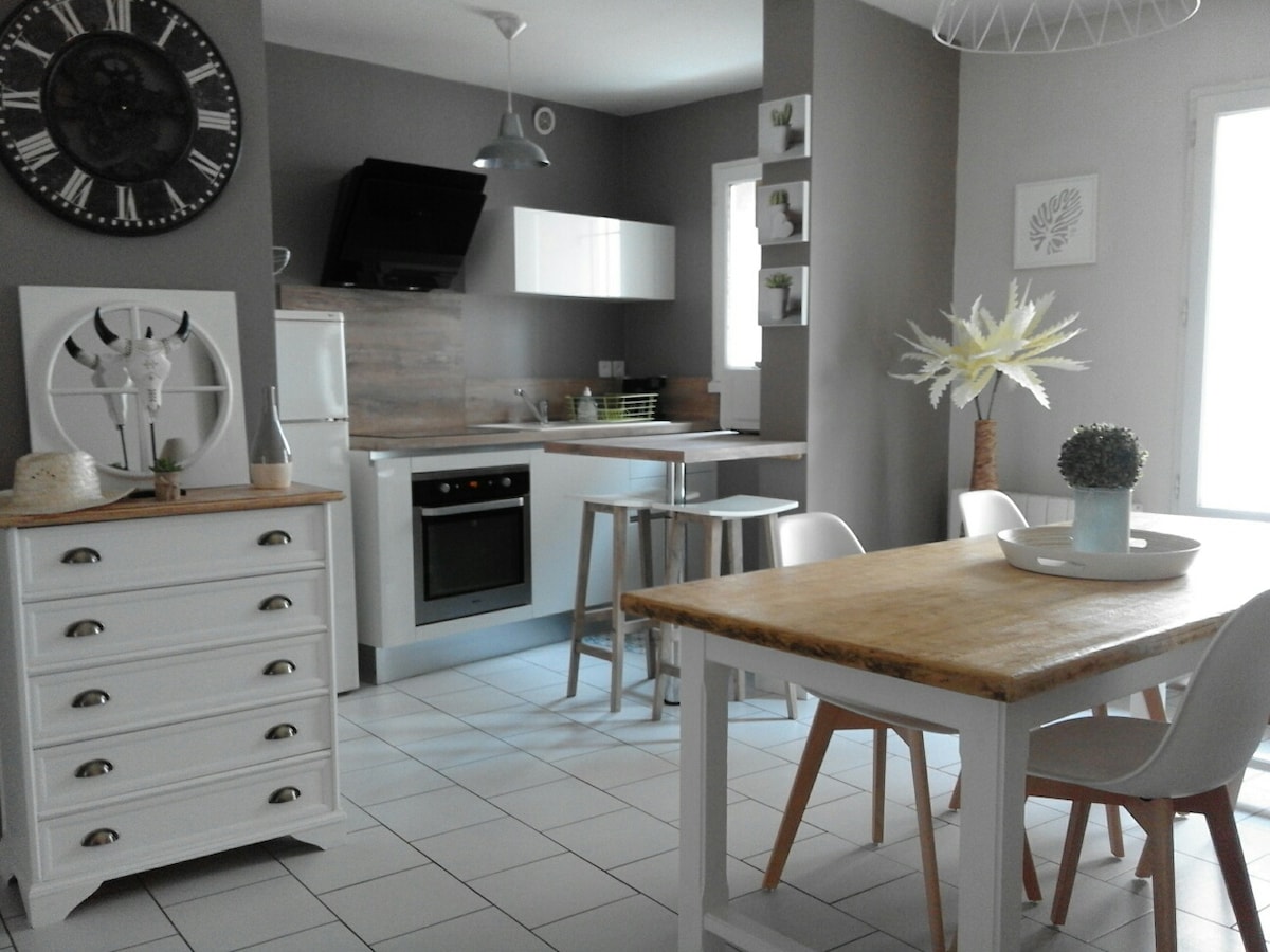 An open kitchen and dining area features modern appliances and a wooden table surrounded by white chairs. Light-colored cabinetry complements the grey walls, while decorative elements such as a clock and potted plants add subtle touches to the space. Natural light enters through the windows.