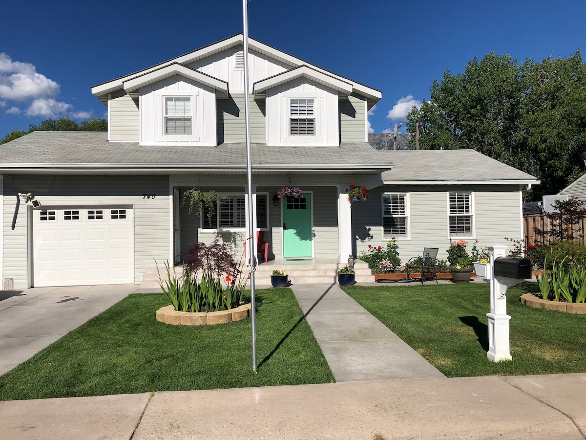 A two-story house is presented, featuring a light-colored exterior with white trim. The front yard is well-maintained, showcasing green grass, flower beds, and vibrant hanging planters. A welcoming mint green door is positioned at the entrance, with a small porch and simple outdoor seating displayed nearby.