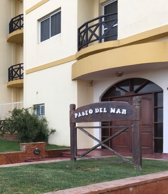 The entrance to the property is marked by a wooden sign reading 'Paseo del Mar.' The building features a modern design with large windows and balconies. A landscaped area with grass and small decorative elements is visible in the foreground.