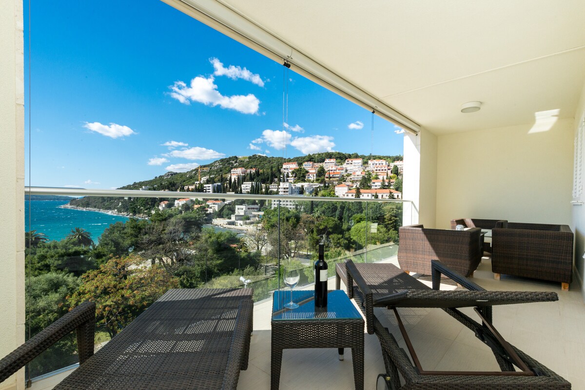 A terrace features modern wicker furniture, including loungers and a small table. A panoramic view of the coastline and hillside is visible, showcasing buildings and greenery under a clear blue sky, with the sea in the background enhancing the tranquil setting.