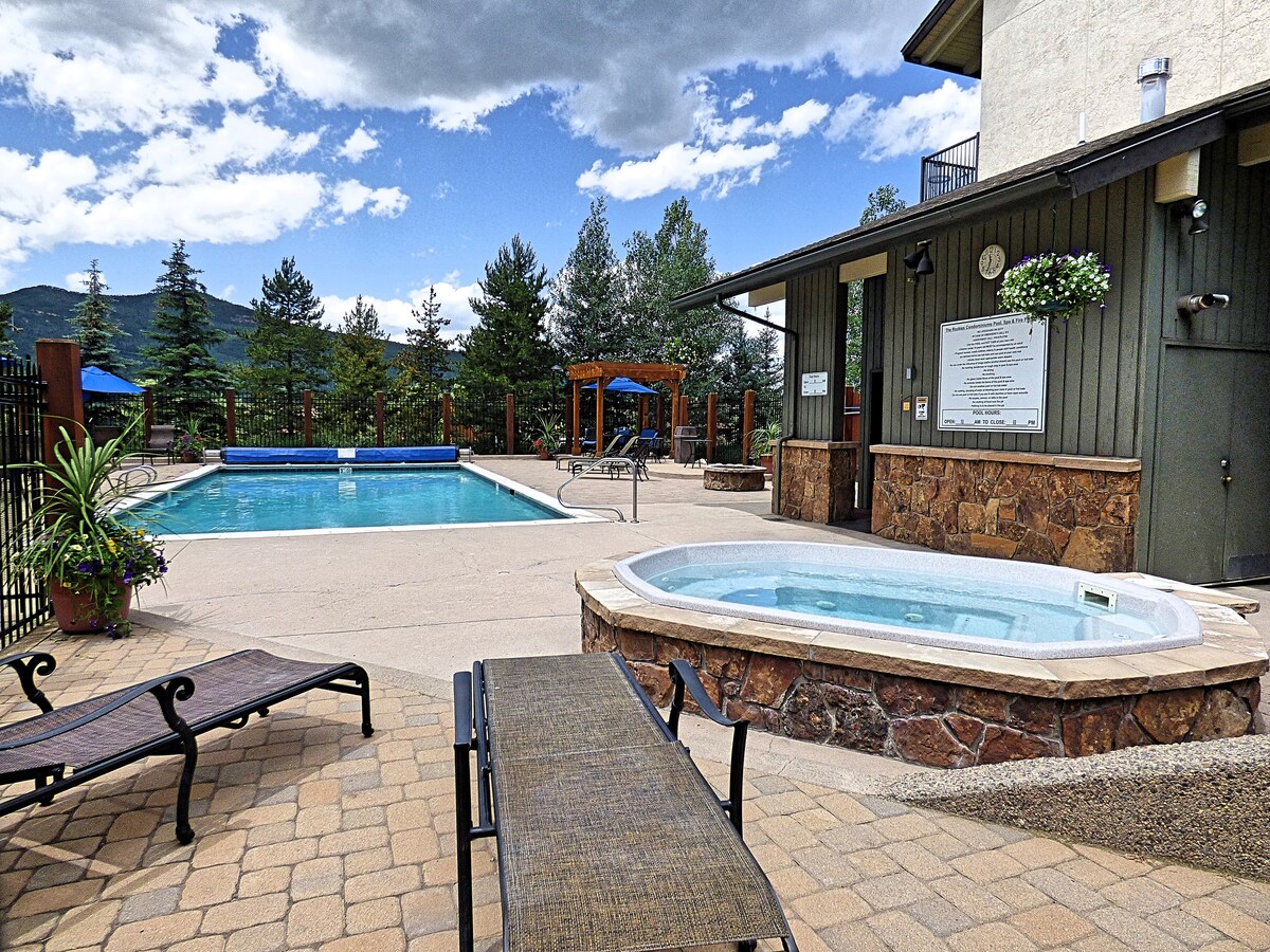 An outdoor pool is situated alongside a hot tub, both surrounded by landscaped areas and mountain views. Lounge chairs are neatly arranged on the patio, inviting relaxation. A wooden gazebo is visible in the background, enhancing the overall leisure atmosphere.