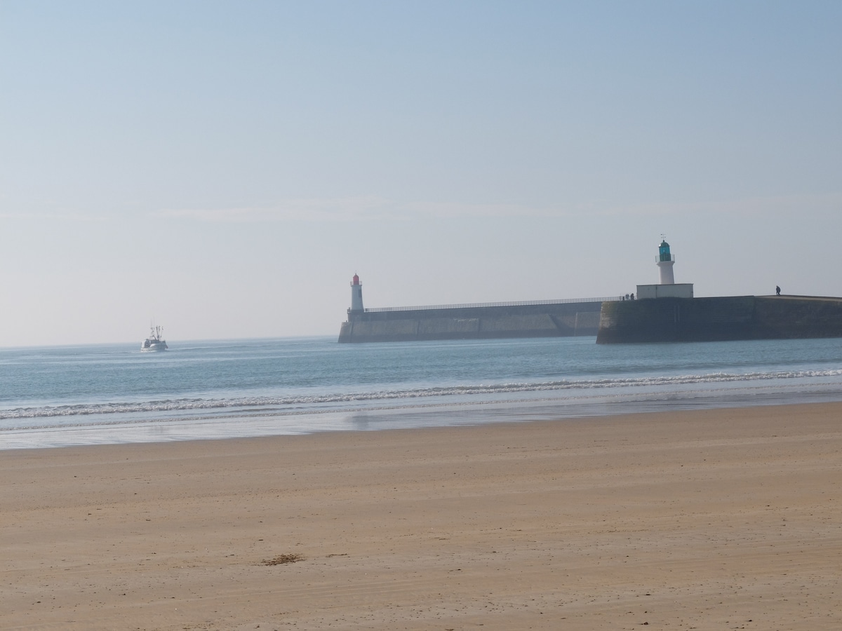 The image captures a serene beach scene with soft sand stretching to calm waters. In the distance, two lighthouses are positioned at the end of a pier, one topped with a red light and the other with a green light. A small boat is seen approaching the pier.