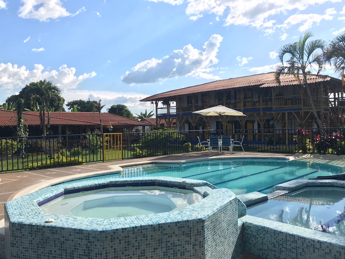 A well-maintained pool area is highlighted, featuring a sparkling blue pool alongside a relaxing jacuzzi. Lush greenery surrounds the space, complemented by colorful flowers and a children's play area. A two-story building provides a charming backdrop, with its traditional architecture and sunny patio.