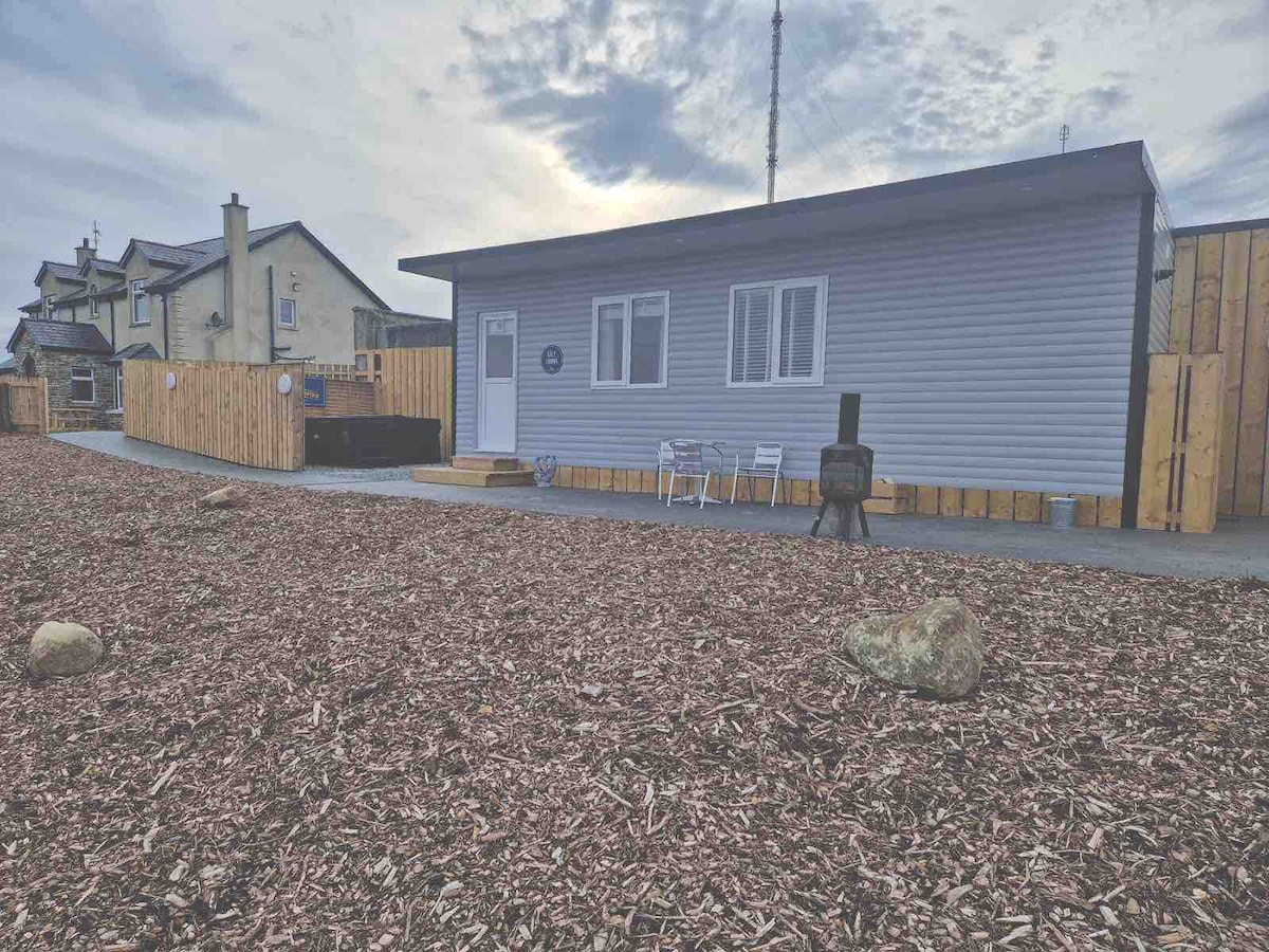A modern lodge features a neutral exterior and multiple windows. A deck is visible with four white chairs, positioned beside a hot tub. The surrounding area consists of a landscaped yard with wood mulch and scattered rocks, enhancing the outdoor space.