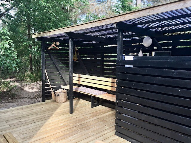 An outdoor bathing area is surrounded by tall trees, featuring a wooden deck and black slatted walls. A bench offers seating, with natural light filtering through the overhead slats. Storage space and hooks for hanging items are visible, enhancing the functional design.