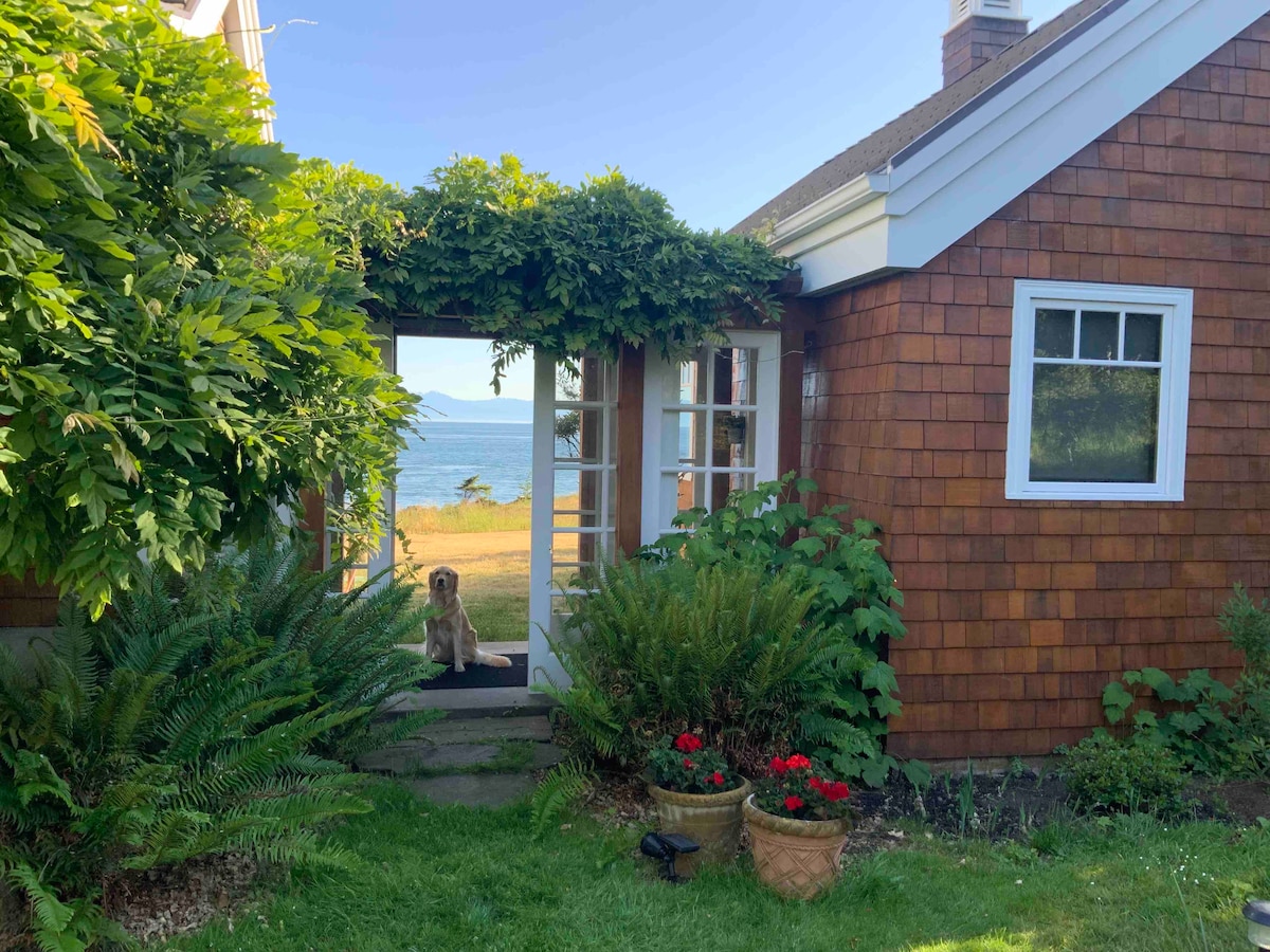 A charming entrance features wooden shingles and large glass doors framed by lush greenery. Potted flowers add vibrant colors at the base, while a scenic view of the water is visible in the background, providing a serene backdrop.