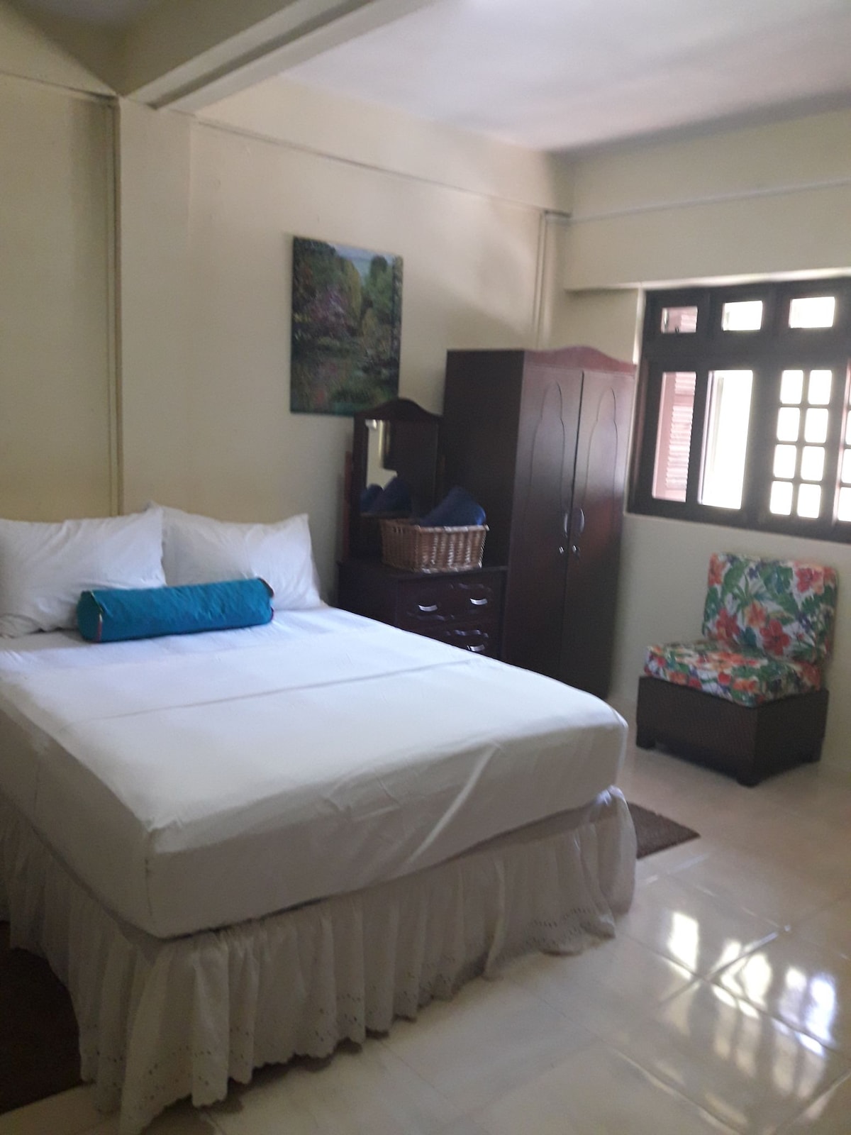 A comfortable sleeping area is presented with a spacious bed dressed in crisp white linens. A colorful bolster pillow is placed on top. A wooden dresser and a flower-patterned chair complement the room, which features soft lighting and a wall-mounted artwork.