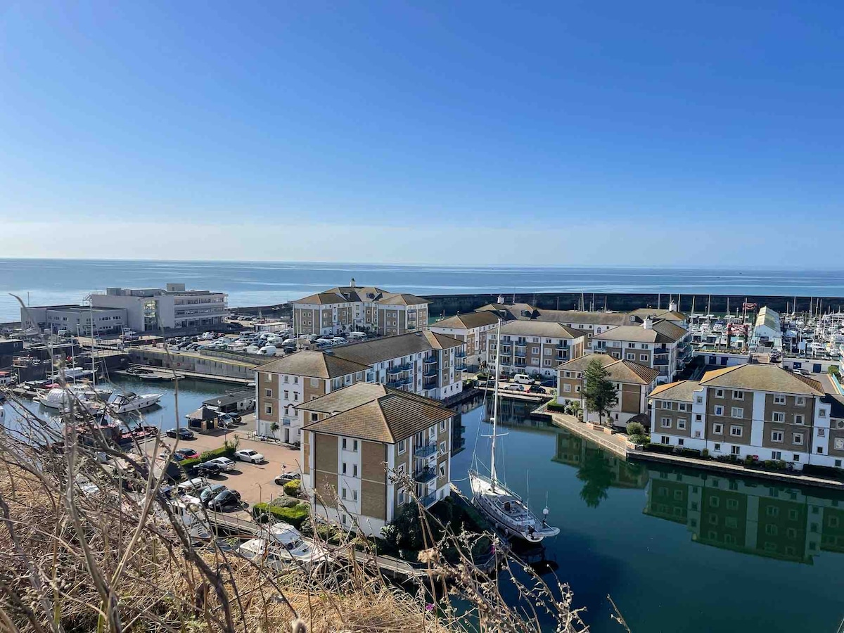 A panoramic view showcases a marina filled with yachts and boats. The surrounding buildings reflect a coastal architectural style, with water gently flowing beside them. The clear blue sky meets the serene sea in the background, emphasizing a tranquil seaside atmosphere.