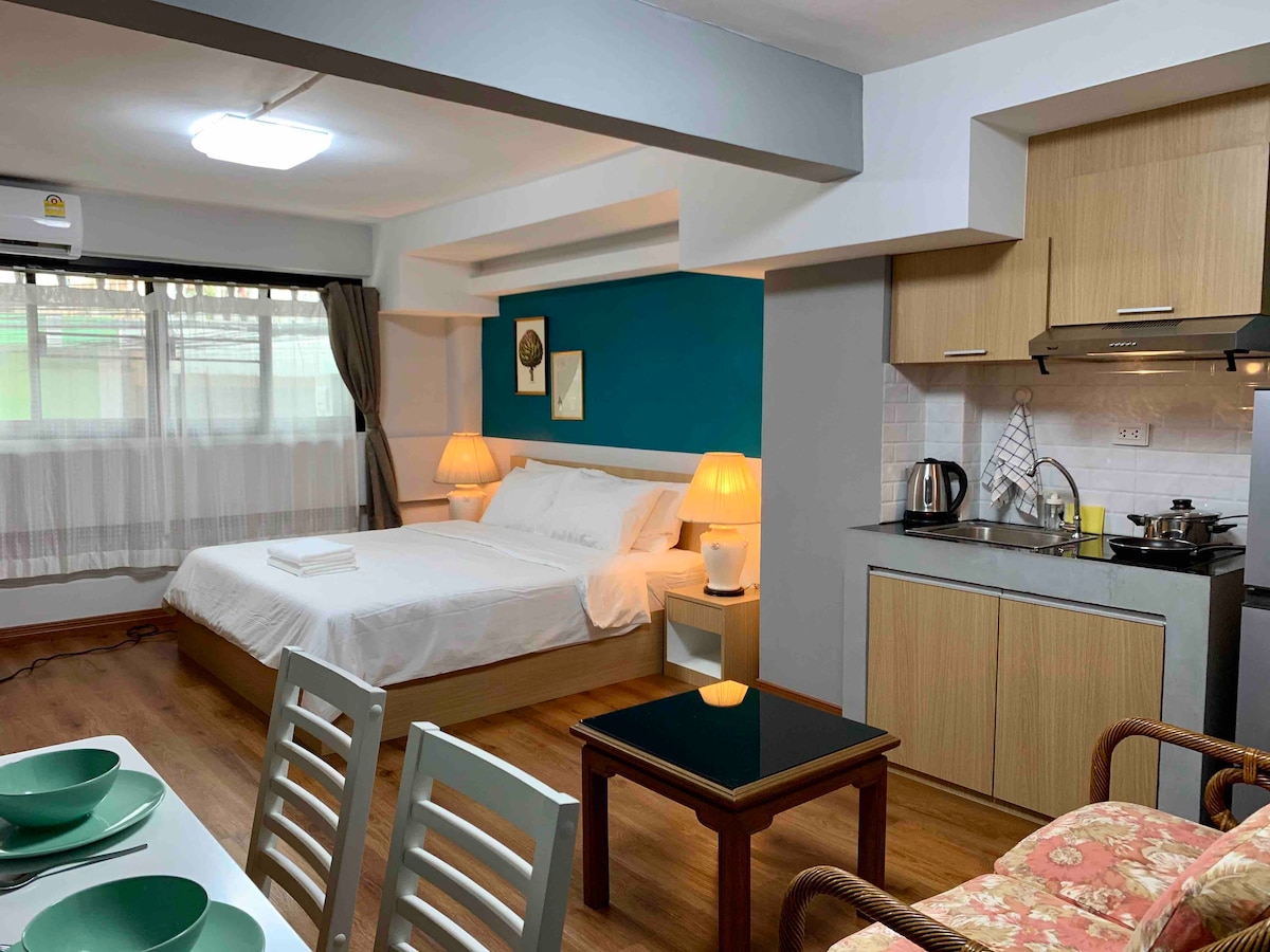 A well-appointed room features a spacious bed framed by soft lighting and a vibrant blue accent wall. A dining area with a small table and four chairs is present, alongside a kitchenette equipped with essential appliances. Large windows provide natural light, complemented by sheer curtains.