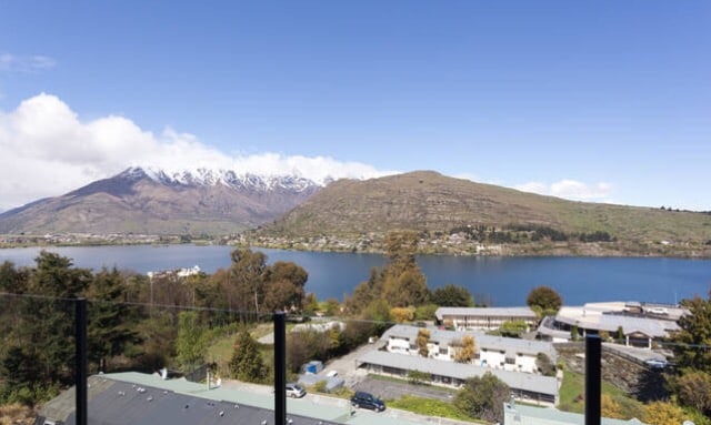 A scenic view encompasses a calm lake bordered by lush greenery and rolling hills. Snow-capped mountains rise majestically in the background, while a clear blue sky complements the serene landscape.