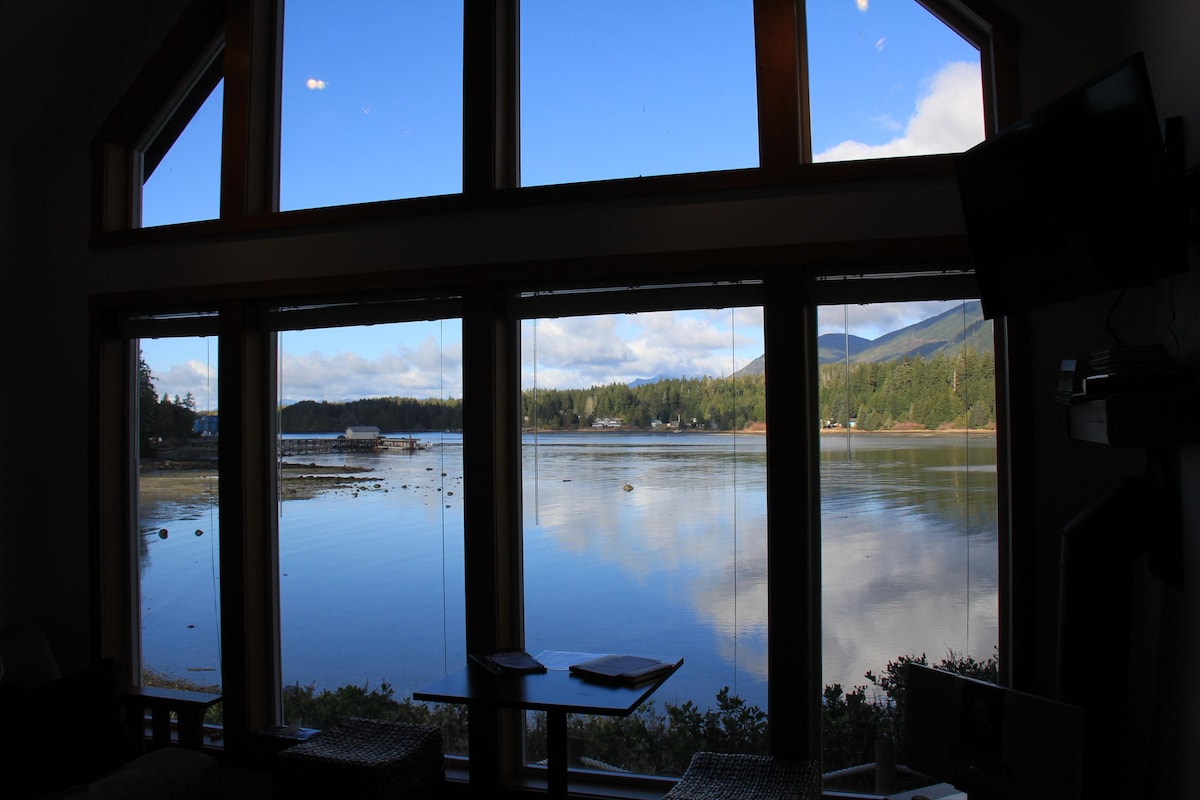 Large windows frame a serene waterfront view, revealing calm waters and surrounding greenery. The open space is illuminated by natural light, emphasizing the tranquil ambiance and the picturesque landscape beyond, where mountains rise gently in the distance.