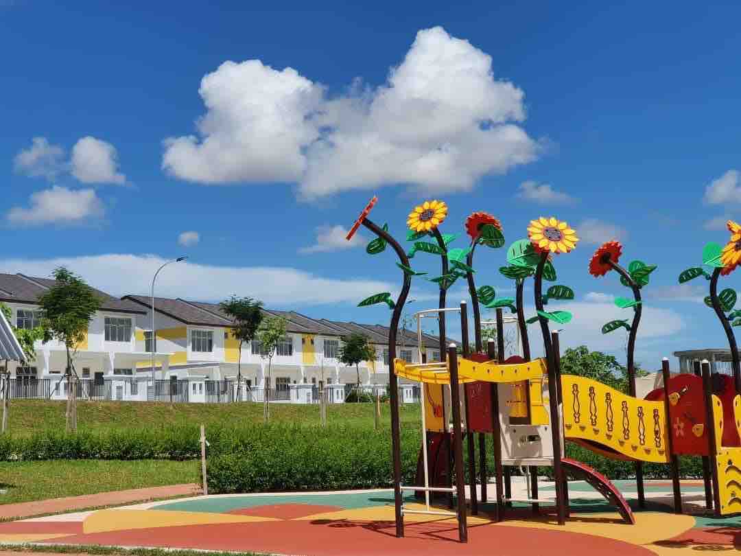 A bright playground is depicted, featuring vibrant flower-themed structures and a colorful sliding area. In the background, the double-storey houses are visible, surrounded by greenery under a clear blue sky with fluffy white clouds.