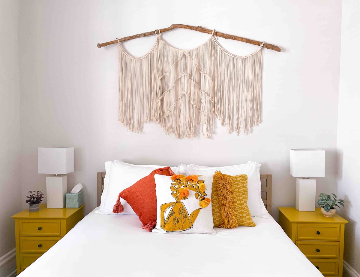 A serene bedroom setting features a spacious bed adorned with bright, decorative pillows. Two yellow nightstands with white lamps flank the bed. Above the bed, a woven wall hanging adds texture to the space, creating a calming atmosphere.