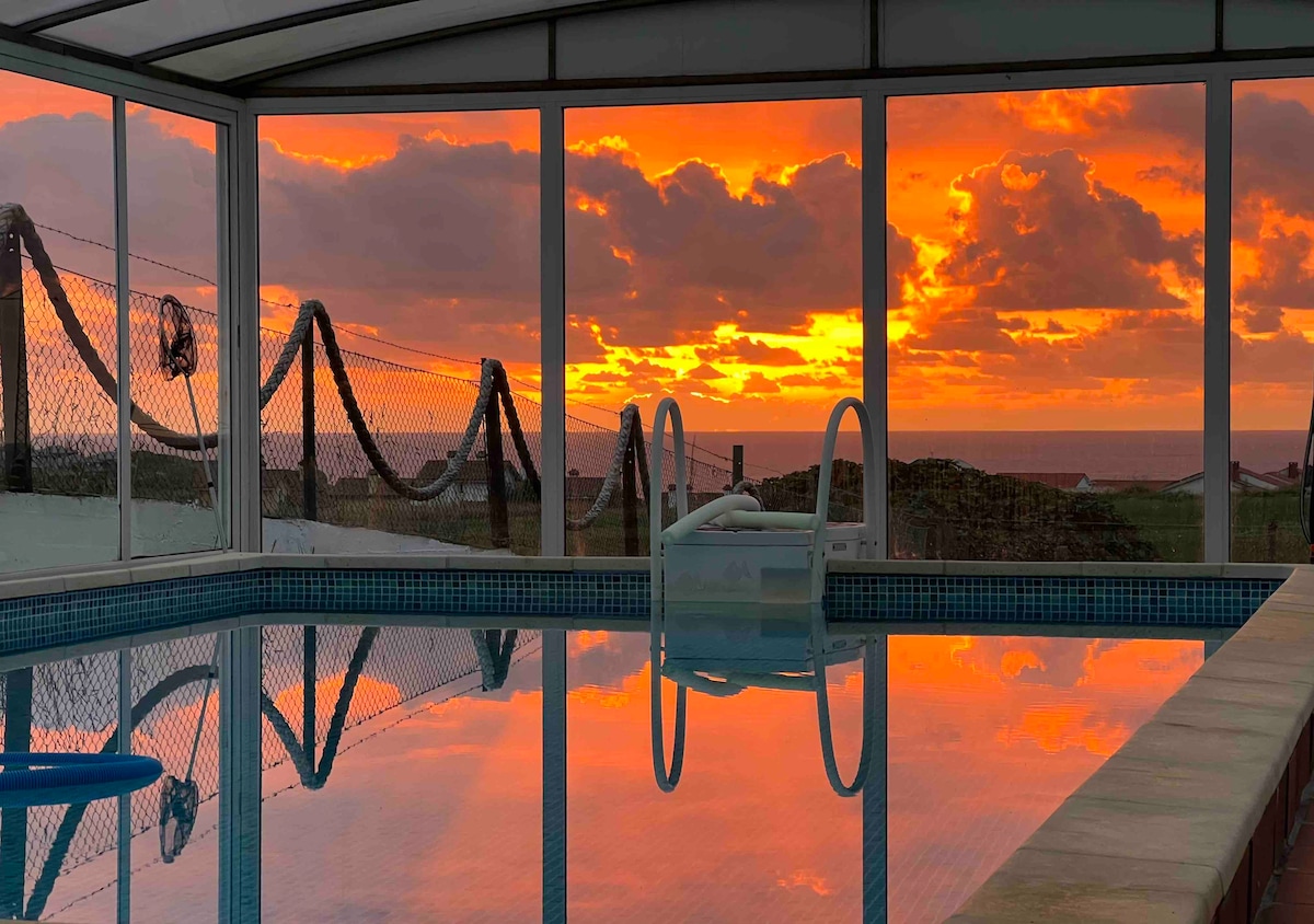 The indoor pool area features large glass walls that provide an unobstructed view of a vibrant sunset over the ocean. The tranquil water reflects the vivid colors of the sky, creating a serene atmosphere. The space is well-lit and enclosed for year-round enjoyment.