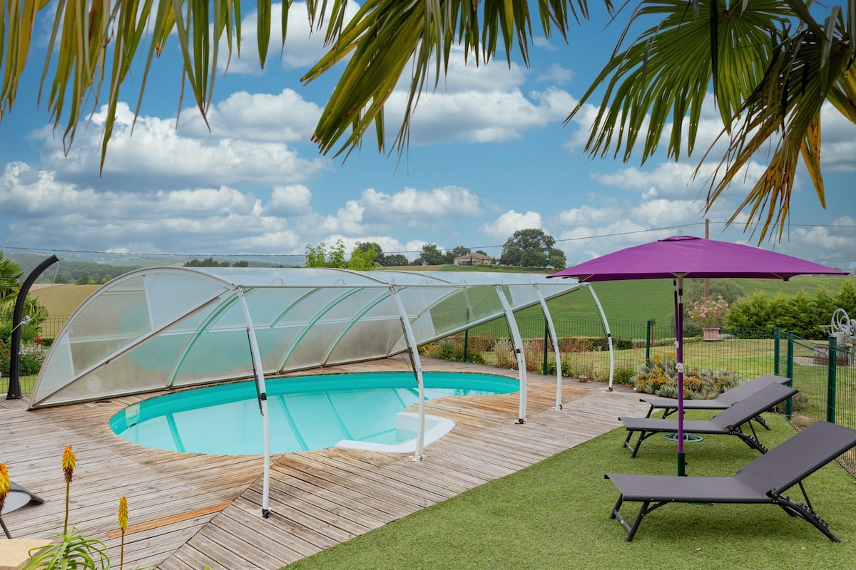 A covered swimming pool is surrounded by a wooden deck and lush greenery. Two lounge chairs are positioned along the pool's edge under a purple umbrella, providing a shaded relaxation area with expansive views of the open countryside in the background.