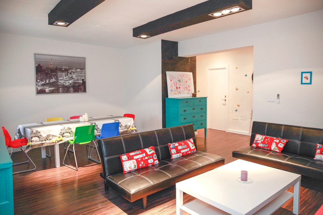 A spacious living area features two sleek black sofas adorned with colorful cushions. A modern white coffee table sits in the center, accompanied by a dining table set for six. Light wood flooring and bright turquoise accents create a welcoming atmosphere.