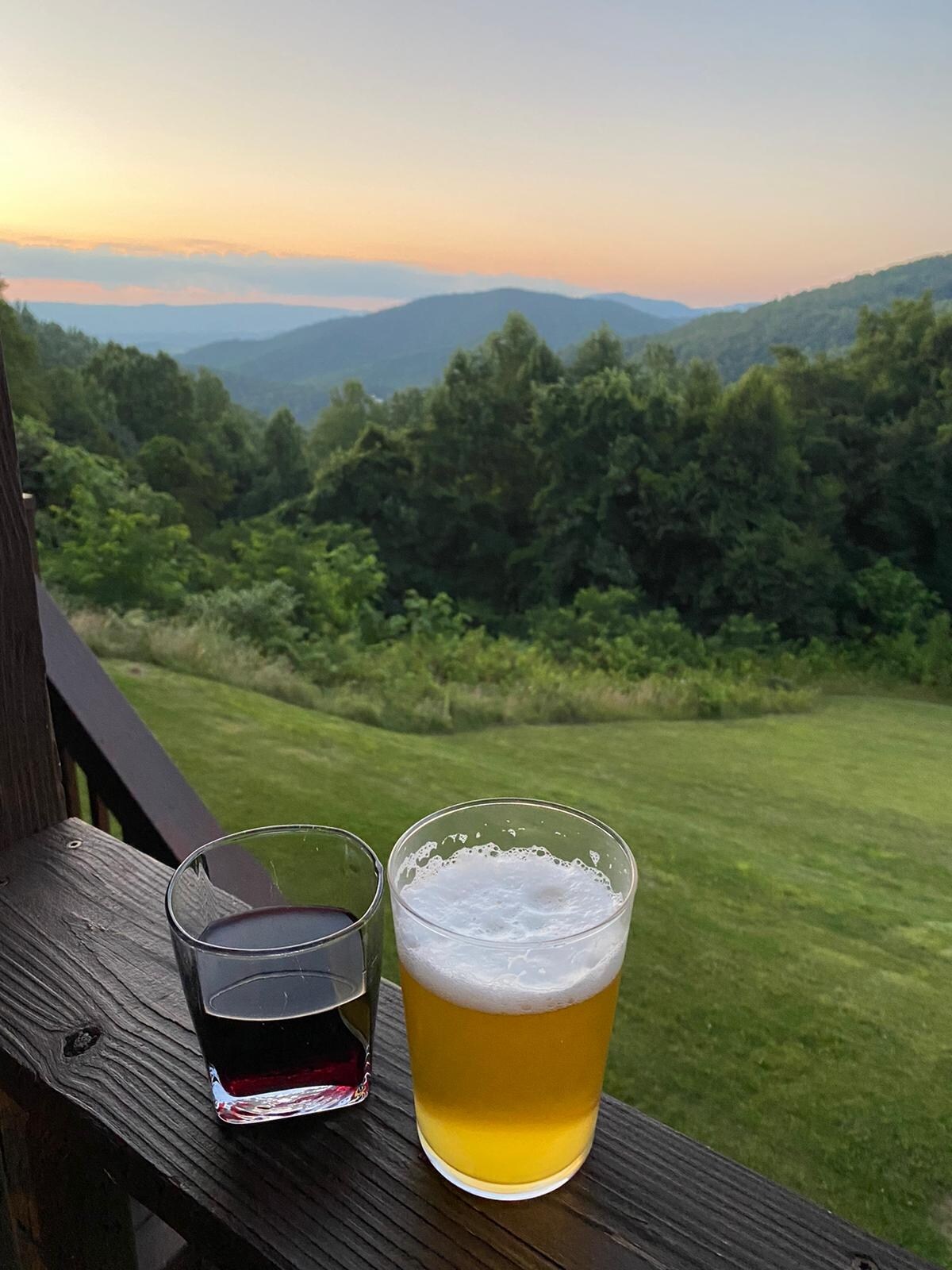 Two glasses sit on a wooden railing, one filled with yellow beer and the other with dark red wine. The background features lush green hills and a soft sunrise illuminating the sky with warm hues, offering a serene mountain view.