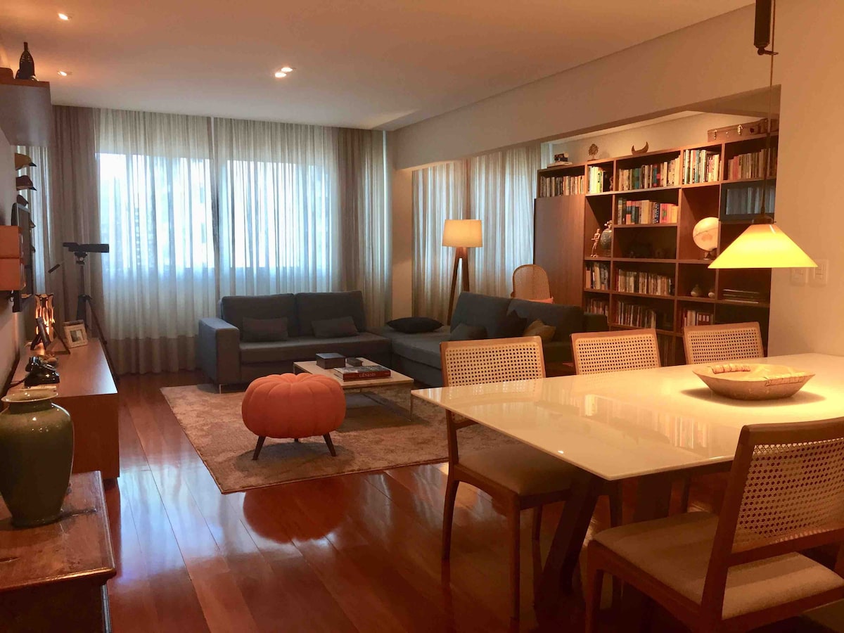 A spacious living area is furnished with two comfortable sofas and a large dining table surrounded by chairs. A bookshelf filled with books is visible, along with decorative items on a side table. Large windows allow natural light to fill the room, which features wooden flooring.