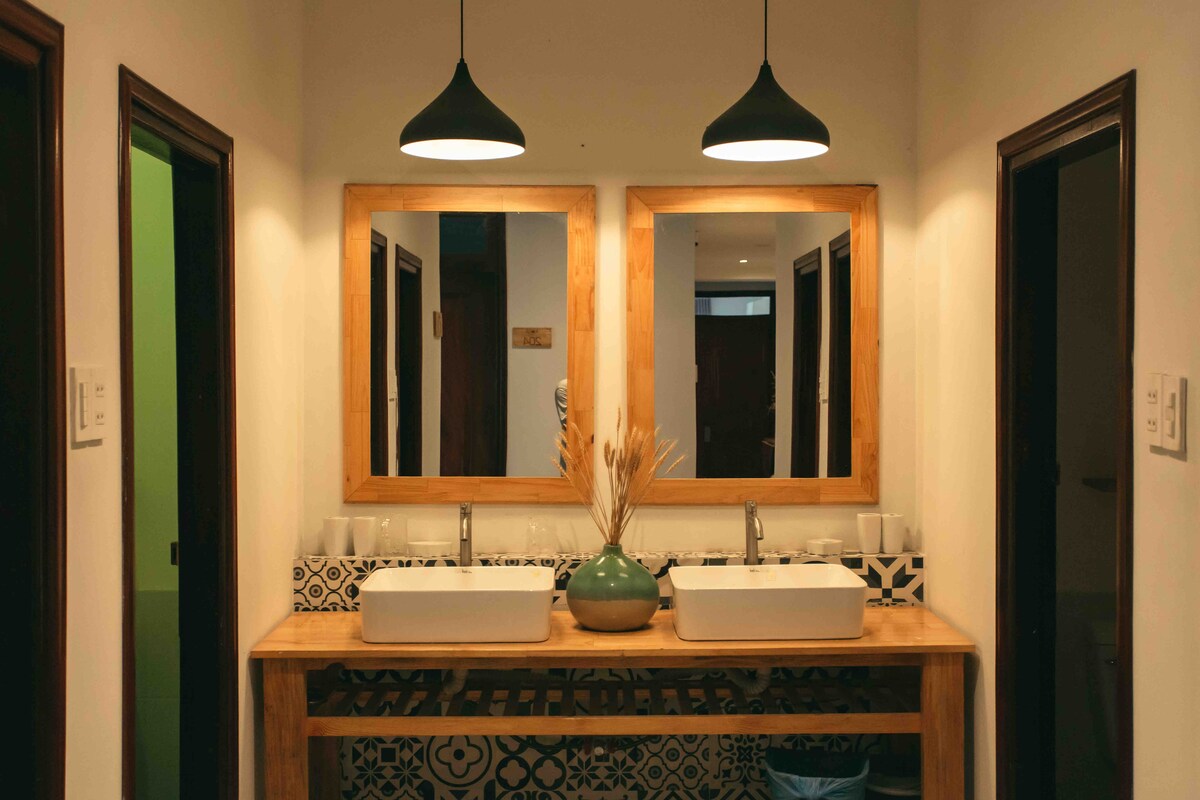 The bathroom features a dual-sink vanity crafted from wood, set against a decorative tiled backsplash. Two large mirrors hang above the sinks, illuminated by stylish pendant lights. Natural elements, such as dried plants in a vase, complete the serene atmosphere.