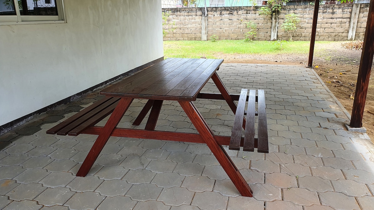 An outdoor covered area features a sturdy wooden picnic table and attached benches, set on a stone-paved surface. The surrounding space is open, with a partially shaded environment, providing a comfortable area for relaxation or meals.