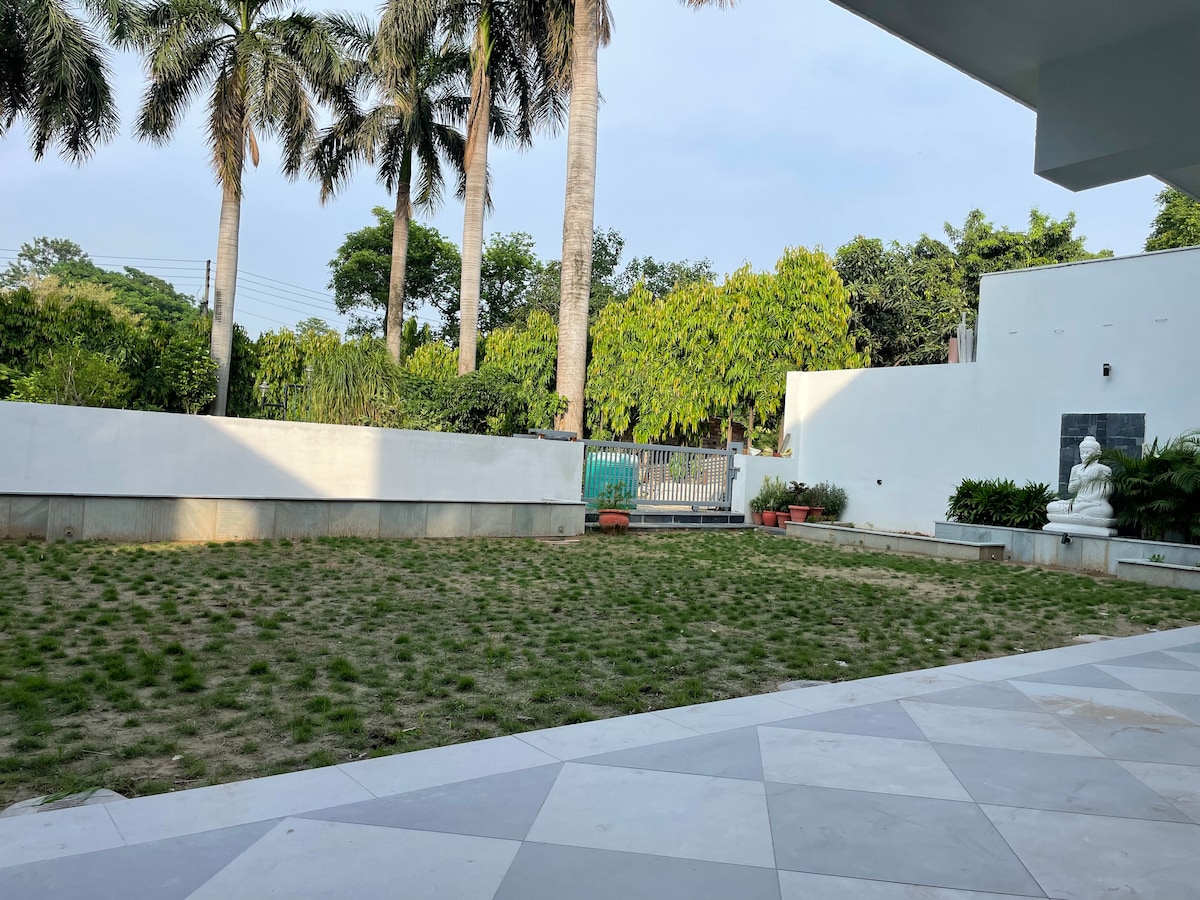 A spacious lawn area is framed by well-maintained greenery and tall palm trees, offering an open and tranquil outdoor space. Neatly arranged flower pots add subtle touches of color against the white walls, enhancing the inviting environment.