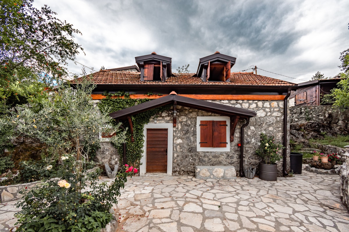 A charming stone exterior of the Little Old House is framed by greenery, with wooden shutters adorning the windows. A cobblestone pathway leads to a wooden door, complemented by flowering plants and well-maintained landscaping, providing a serene and inviting entryway.