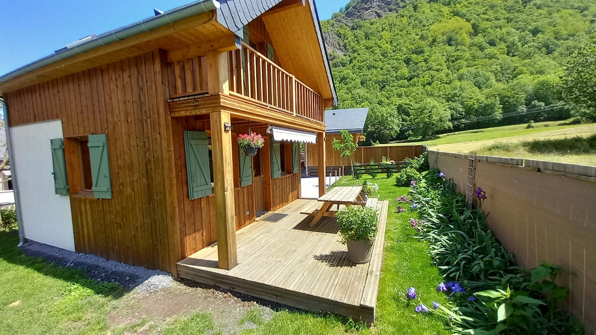 The wooden chalet features a spacious terrace with a wooden table and potted plants. Lush greenery surrounds the area, including flowers along the edge of the garden. The building displays traditional shutters and is set against a backdrop of rolling hills covered in trees.