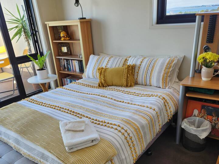 A cozy bedroom with a striped yellow and white bedspread, accented by a matching decorative pillow. A small wooden bookshelf is beside the bed, while a desk holds a vase of yellow flowers. The room opens to a balcony with outdoor seating and a view of the ocean in the distance