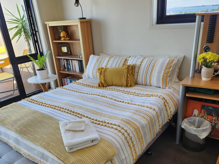 A cozy bedroom with a striped yellow and white bedspread, accented by a matching decorative pillow. A small wooden bookshelf is beside the bed, while a desk holds a vase of yellow flowers. The room opens to a balcony with outdoor seating and a view of the ocean in the distance