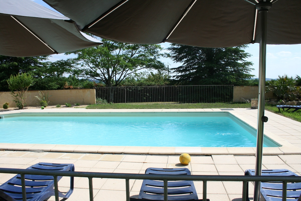 A tranquil outdoor area features a private swimming pool surrounded by a paved deck. Sun loungers are positioned nearby, complemented by large umbrellas providing shade. Lush greenery and a low fence offer a natural backdrop, enhancing the peaceful atmosphere of the space.