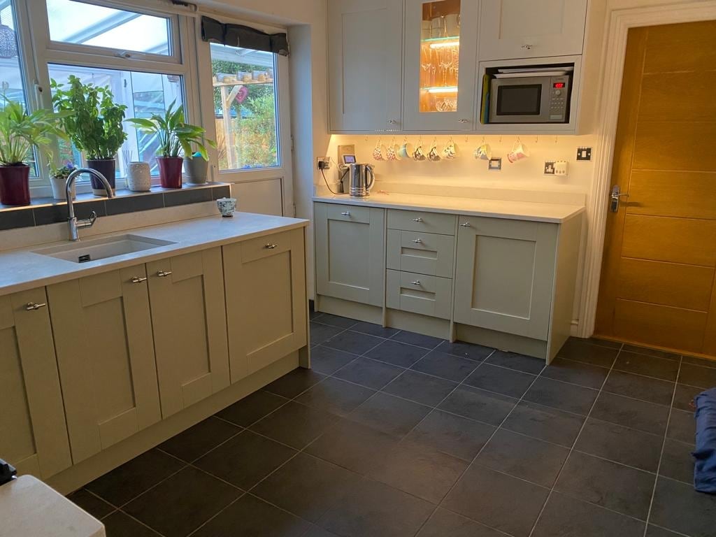 The kitchen features modern cabinetry in a muted color, complemented by a spacious counter area and a stainless steel microwave. Natural light enters through the windows adorned with potted plants, and dark tiles cover the floor, creating a functional cooking space.