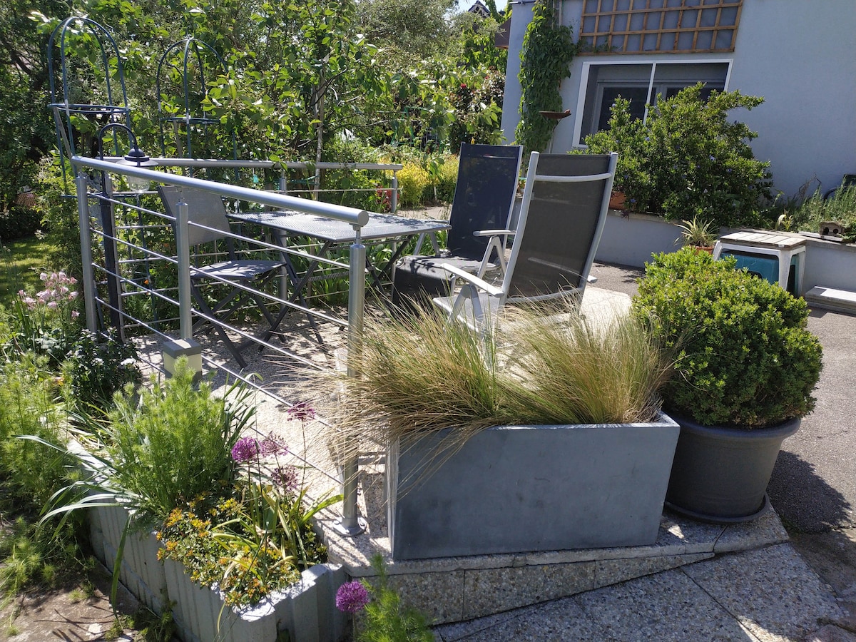A quiet outdoor terrace is surrounded by lush greenery and colorful flowers. A small metal table and two chairs are positioned, offering a serene space for relaxation. Decorative planters filled with grass and blooming plants enhance the inviting atmosphere.