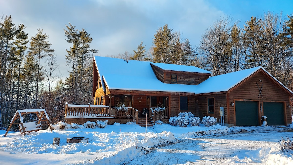 A spacious wooden cabin is set against a snowy landscape, featuring a covered porch with a swing. The exterior is adorned with a combination of green doors and natural wood accents, while surrounding trees rise gently in the background under a clear blue sky.