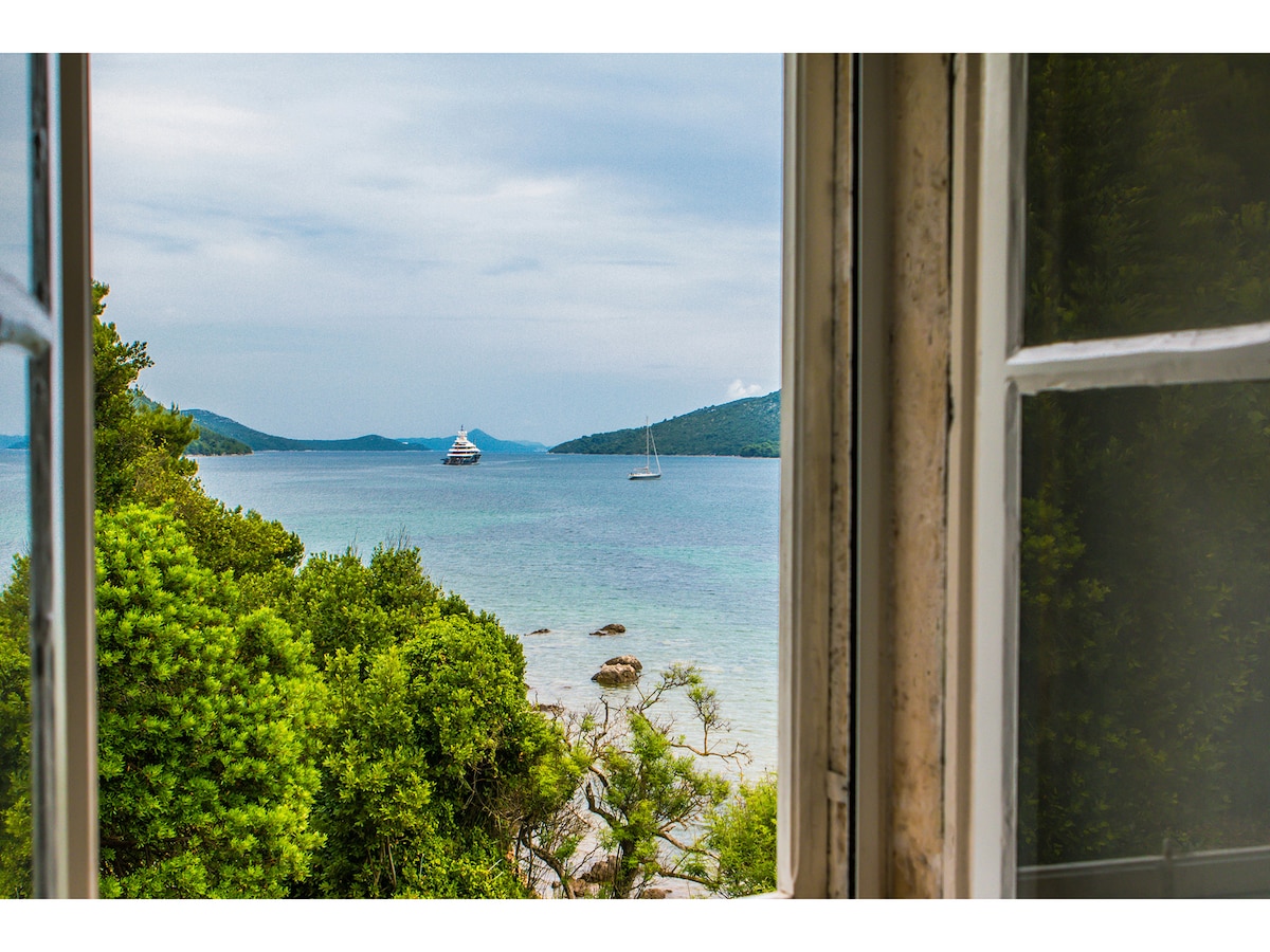 A view from an open window reveals a serene seascape, featuring calm waters and distant sailboats anchored amidst lush greenery. Rolling hills create a picturesque backdrop, enhancing the natural beauty of the coastline.