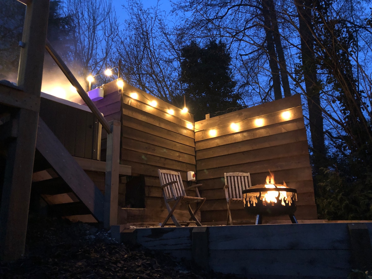 A relaxing outdoor space is illuminated by string lights, featuring a fire pit surrounded by two wooden chairs. A hot tub is partially visible in the background, set against a backdrop of trees, creating a tranquil evening ambiance.