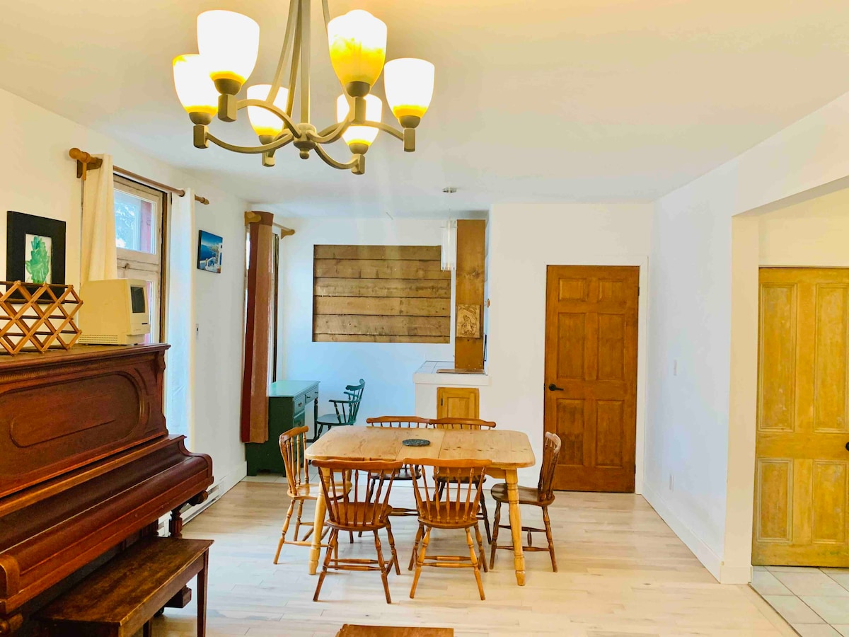 A dining area is visible, featuring a wooden table surrounded by several chairs. Soft lighting is provided by a chandelier above. A vintage piano stands against one wall, complemented by wooden accents. Natural light enters through windows, enhancing the inviting space.