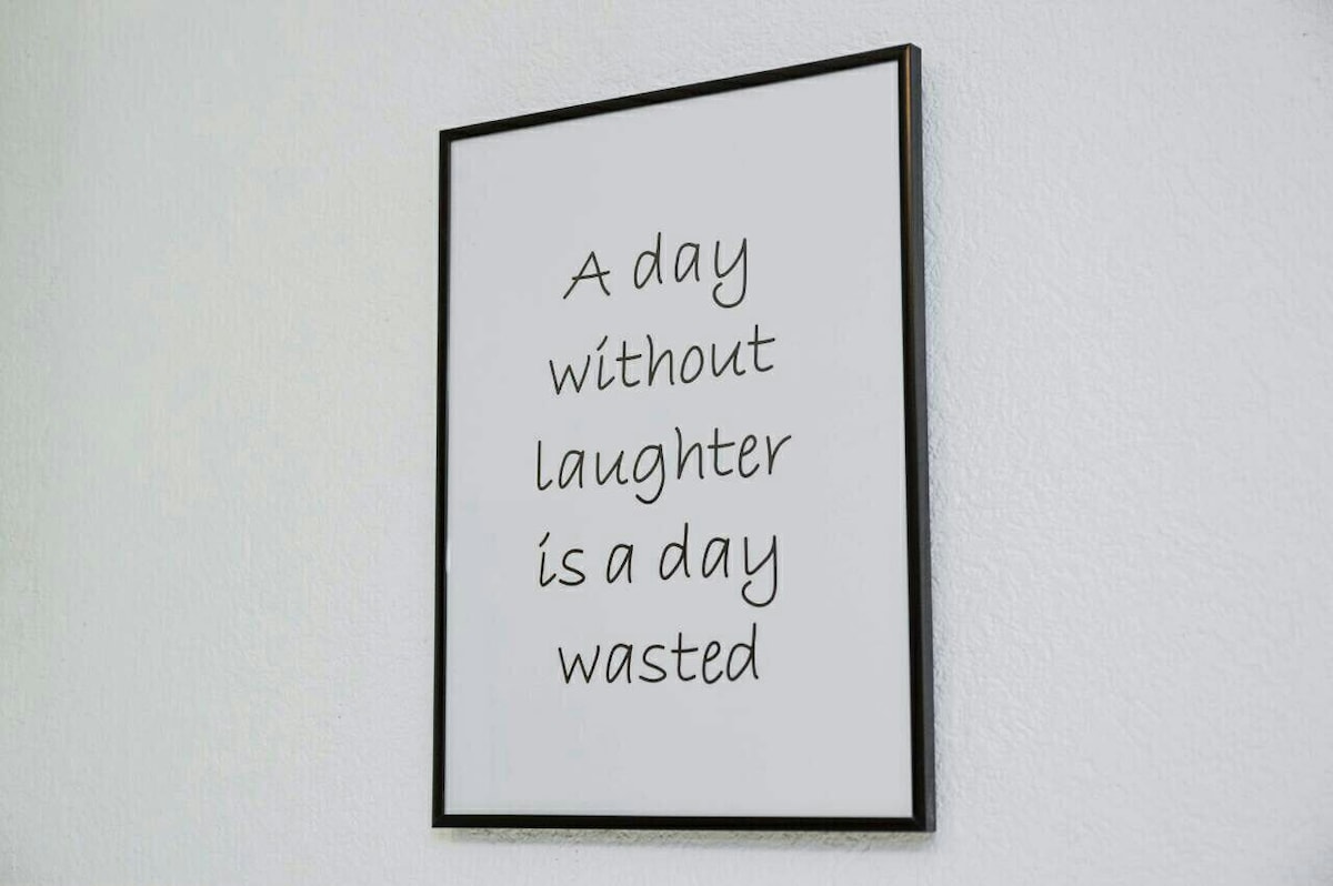 A framed quote is displayed on a light-colored wall. The text, written in a decorative font, reads 'A day without laughter is a day wasted.' The frame is simple and black, providing a contrast to the wall.
