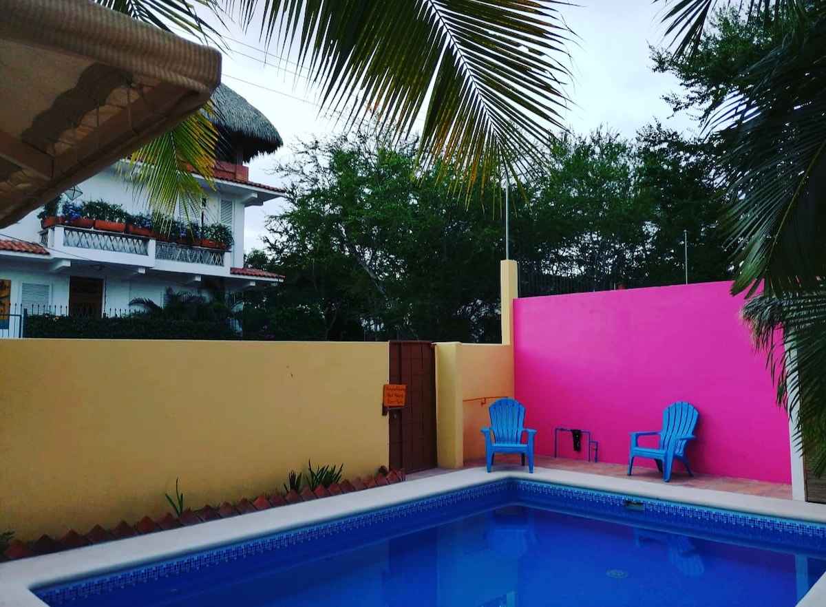 A tranquil swimming pool is framed by a colorful pink wall and lush greenery. Two blue chairs are positioned nearby, inviting relaxation. A shaded area is visible in the top corner, providing comfort while enjoying the outdoor space.