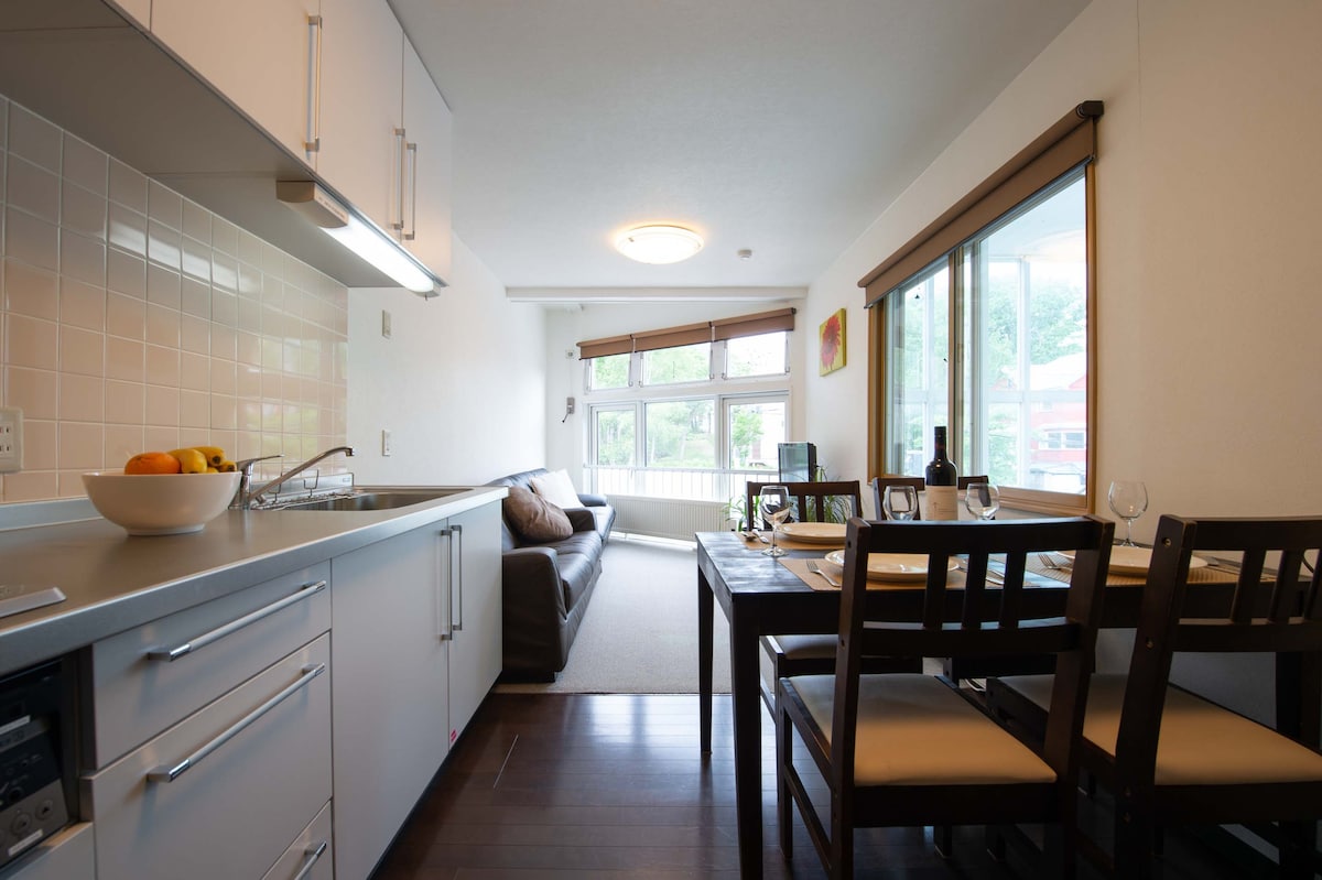 A modern kitchen area is visible, accompanied by a dining table set for four. A comfortable sofa is positioned nearby, with large windows allowing natural light to fill the space. The flooring is dark wood, contrasting with the light-colored walls.