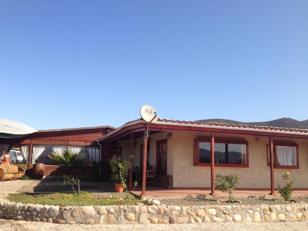 A single-story exterior presents a warm façade with large windows framed in wood. A porch is visible, complemented by potted plants. The surrounding landscape exhibits a sunny, clear sky, and the building is partially shaded by a nearby roof structure.