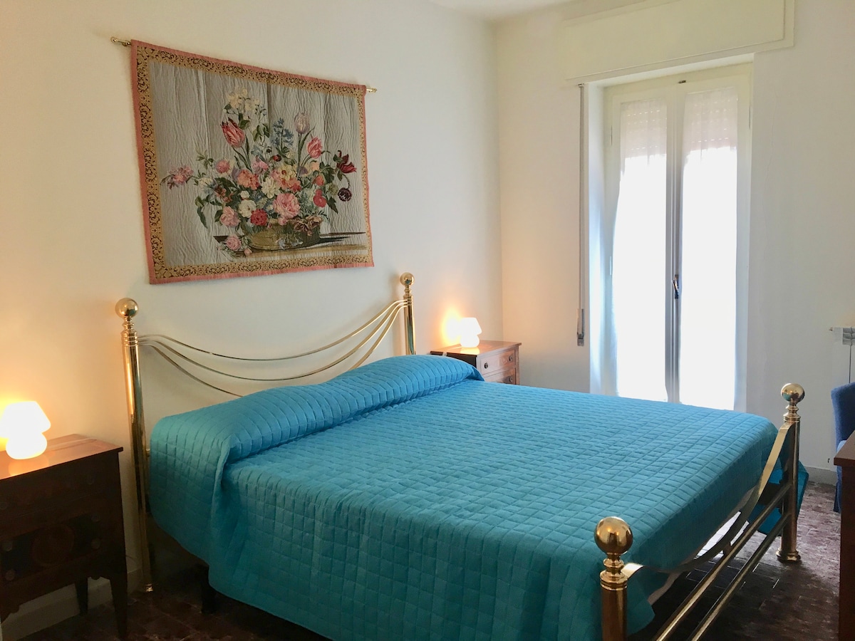 A well-lit bedroom features a spacious bed with a turquoise quilt. Two wooden nightstands are placed on either side of the bed, topped with soft glowing lamps. A floral textile hangs on the wall above the bed, and natural light enters through a pair of curtains.