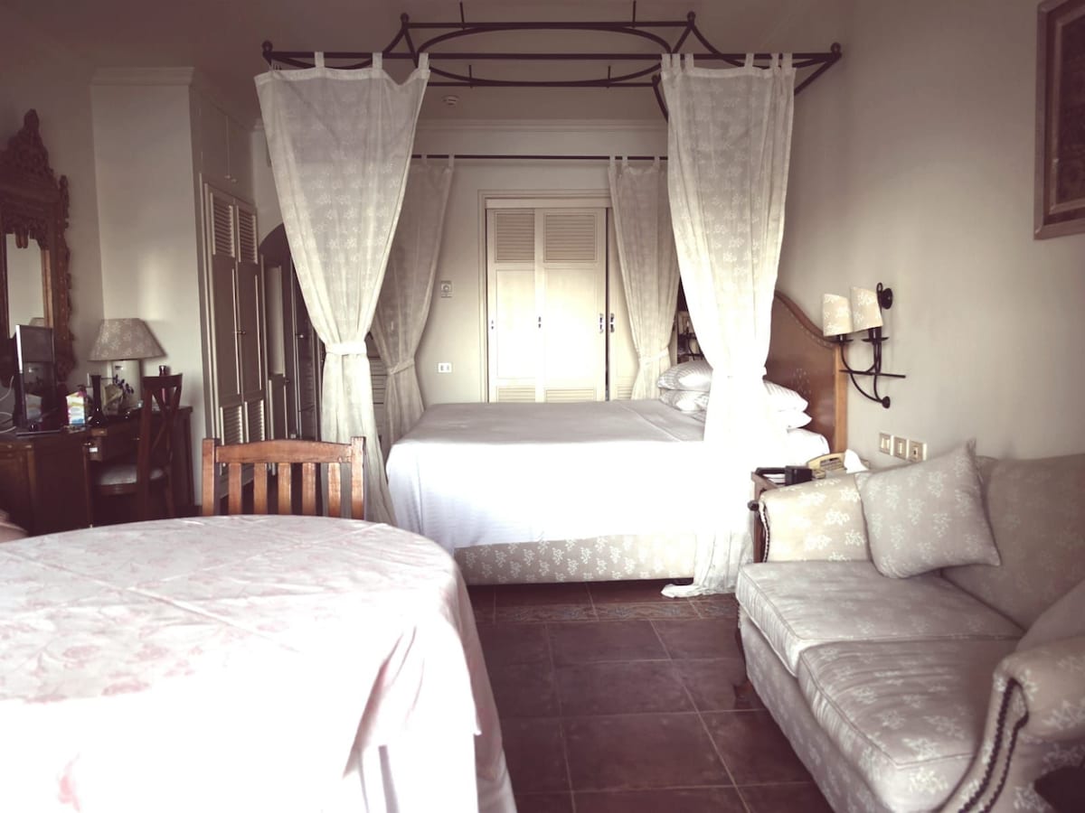 A comfortable room features a spacious bed draped in soft linens, surrounded by flowing curtains. A small dining table is positioned nearby, accompanied by a wooden chair. A cozy seating area includes a patterned sofa, complementing the elegant décor.