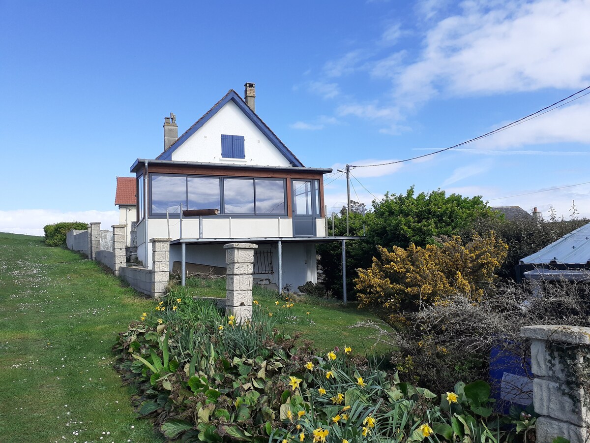 A two-story house with a charming facade is set against a clear blue sky. A spacious veranda with large windows offers views of the surrounding greenery and flowering plants. The exterior features a well-maintained lawn and stone walls, enhancing the home's appeal.