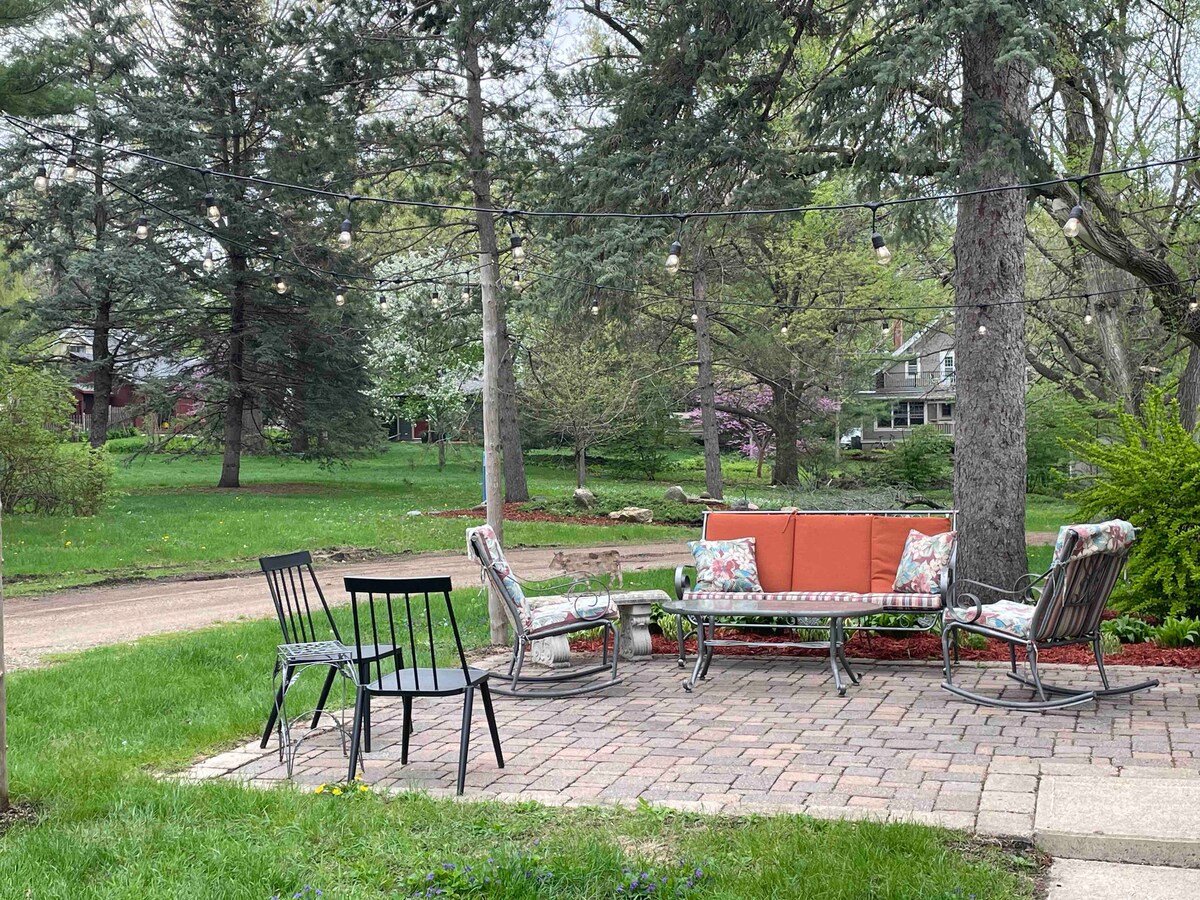 A patio area features a circular table surrounded by cushioned chairs with floral patterns. Soft string lights hang above, adding a gentle illumination. The surrounding greenery includes trees and shrubs, creating a peaceful outdoor setting.