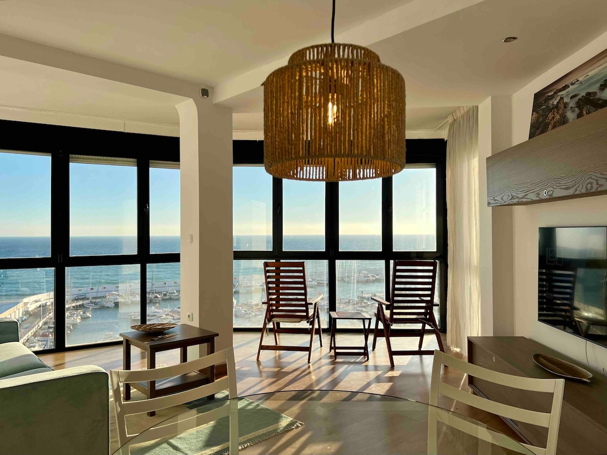 An inviting living area is highlighted by large windows offering unobstructed views of the Mediterranean Sea and Marbella sports harbor. Two wooden lounge chairs are positioned for relaxation, while a stylish rattan light fixture adds character to the room. A soft-colored sofa and a small glass table complete the setup.
