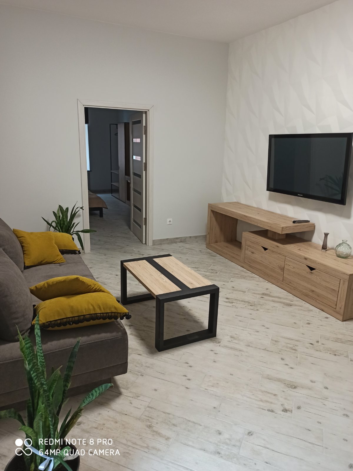 A modern living area features a grey sofa with yellow cushions, complemented by a minimalistic black coffee table. A light wooden media unit is mounted against the wall, housing a flat-screen television. A potted plant adds a touch of greenery to the space.